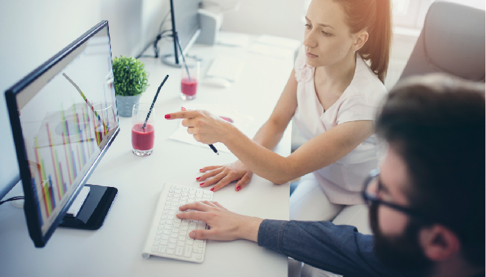 Two coworkers sit together and point at a computer screen