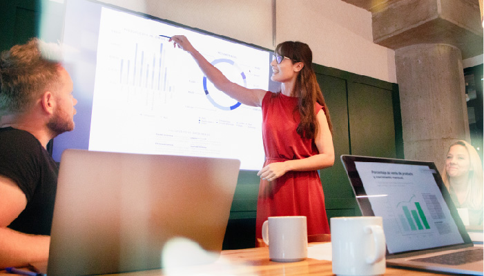 A woman gives a presentation in an office conference room and points at a bar graph displayed on a large screen