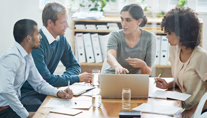 A woman points at her laptop screen while her three co-workers  listen and take notes