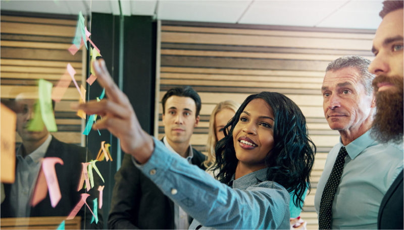 A woman points at a wall full of sticky notes as her coworkers look on