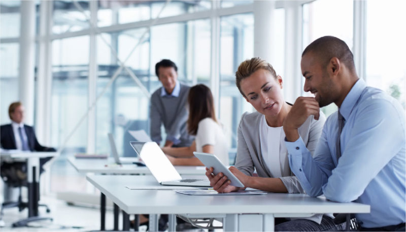 A male coworker and female coworker sit together at a desk in the office and look at an electronic tablet