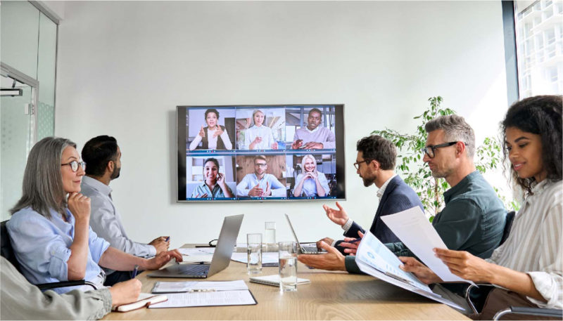 A group of coworkers sit in a conference room and speak with remote colleagues who appear via Zoom on a large screen