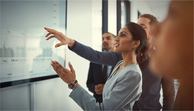 A woman and her coworkers look at a large monitor that shows bar graphs