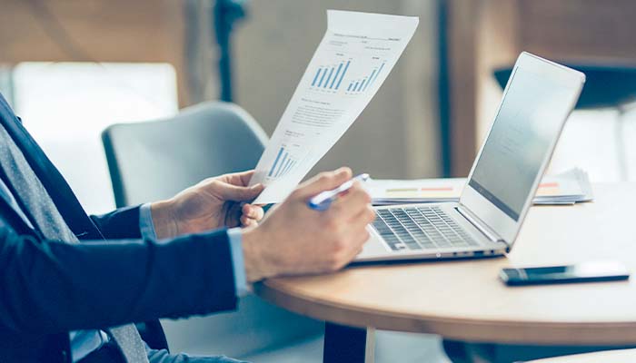 A man sits at a table with his laptop open while he looks at paper with graphs printed on it