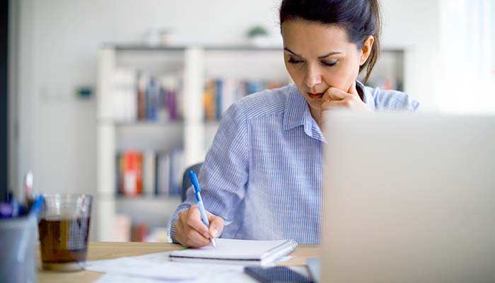 A woman writing down notes while sitting at her desk with her laptop open