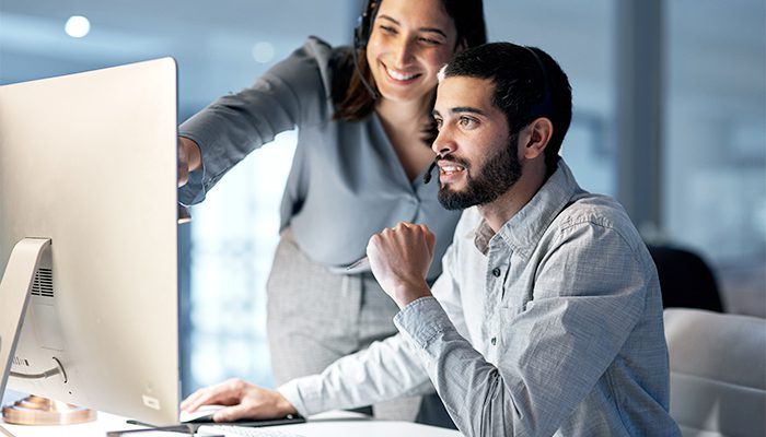 A female co-worker stands and points at the computer screen of her male co-worker who is sitting at his desk