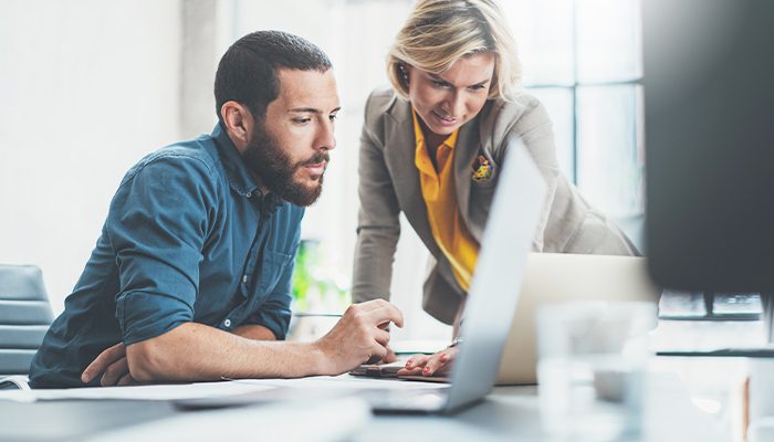 A man and woman work together on their laptops at a desk in the office